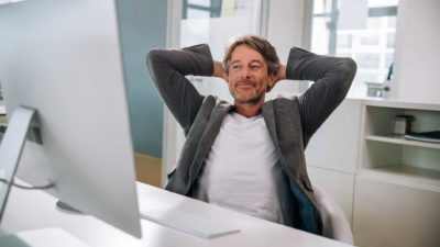 Contented looking man leans back in his chair at his desk and smiles.