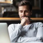 A male investor sits at his desk looking at his laptop screen holding his hand to his chin pondering whether to buy Macquarie shares