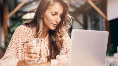 A woman sits in a cafe wearing a polka dotted shirt and holding a latte in one hand while reading something on a laptop that is sitting on the table in front of her