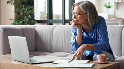 A woman sits in her home with chin resting on her hand and looking at her laptop computer with some reflection with an assortment of books and documents on her table.