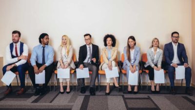 a line up of job interview candidates sit in chairs against a wall clutching CVs on paper in an office setting.