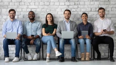 a line of job applicants sit on stools against a brick wall in an office environment, various holding laptops , devices and paper, as though waiting to be interviewed for a position.