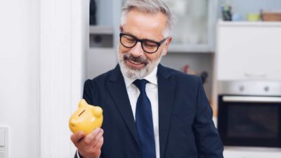 A man in a suit smiles at the yellow piggy bank he holds in his hand.