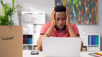 A man sits in despair at his computer with his hands either side of his head, staring into the screen with a pained and anguished look on his face, in a home office setting.