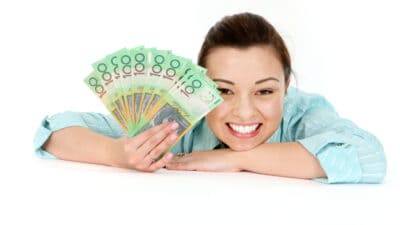 Smiling woman with her head and arm on a desk holding $100 notes out, symbolising dividends.