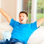 Man raising both his arms in the air with a piggy bank on his lap, symbolising a record high.