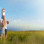 Three small children reach up to hold a toy rocket high above their heads in a green field with a blue sky above them.