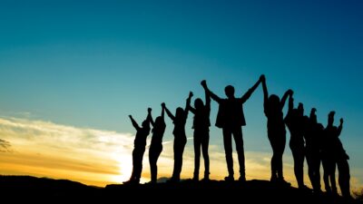 The silhouettes of ten people holding hands with their arms raised against the sky, as the sun rises or sets in the background.