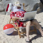 A fun depiction of summer Santa Claus -- wearing red swimming trunks and Hawaiian shirt -- sitting in a deck chair on his laptop at the beach.