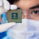 A tech worker wearing a mask holds computer chip up to the camera.