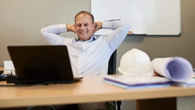 Cheerful businessman with a mining hat on the table sitting back with his arms behind his head while looking at his laptop's screen.