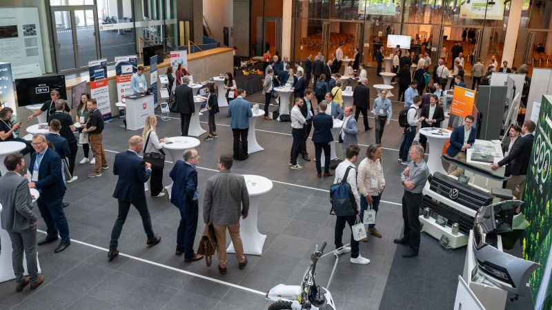 Das Bild zeigt eine Messehalle mit Blick von oben auf Stehtische, Stände und Besucher, die im Gespräch miteinander und mit Ausstellern sind.