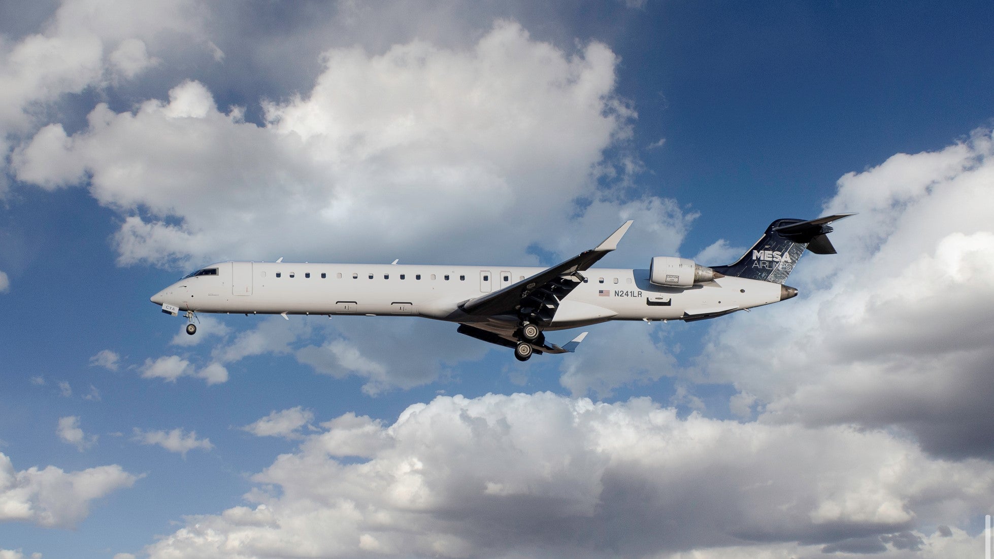 A white Mesa regional jet with a blue tail flying through blue sky and clouds as it comes in for a landing. The FAA's new rules allow planes to descend more efficiently.