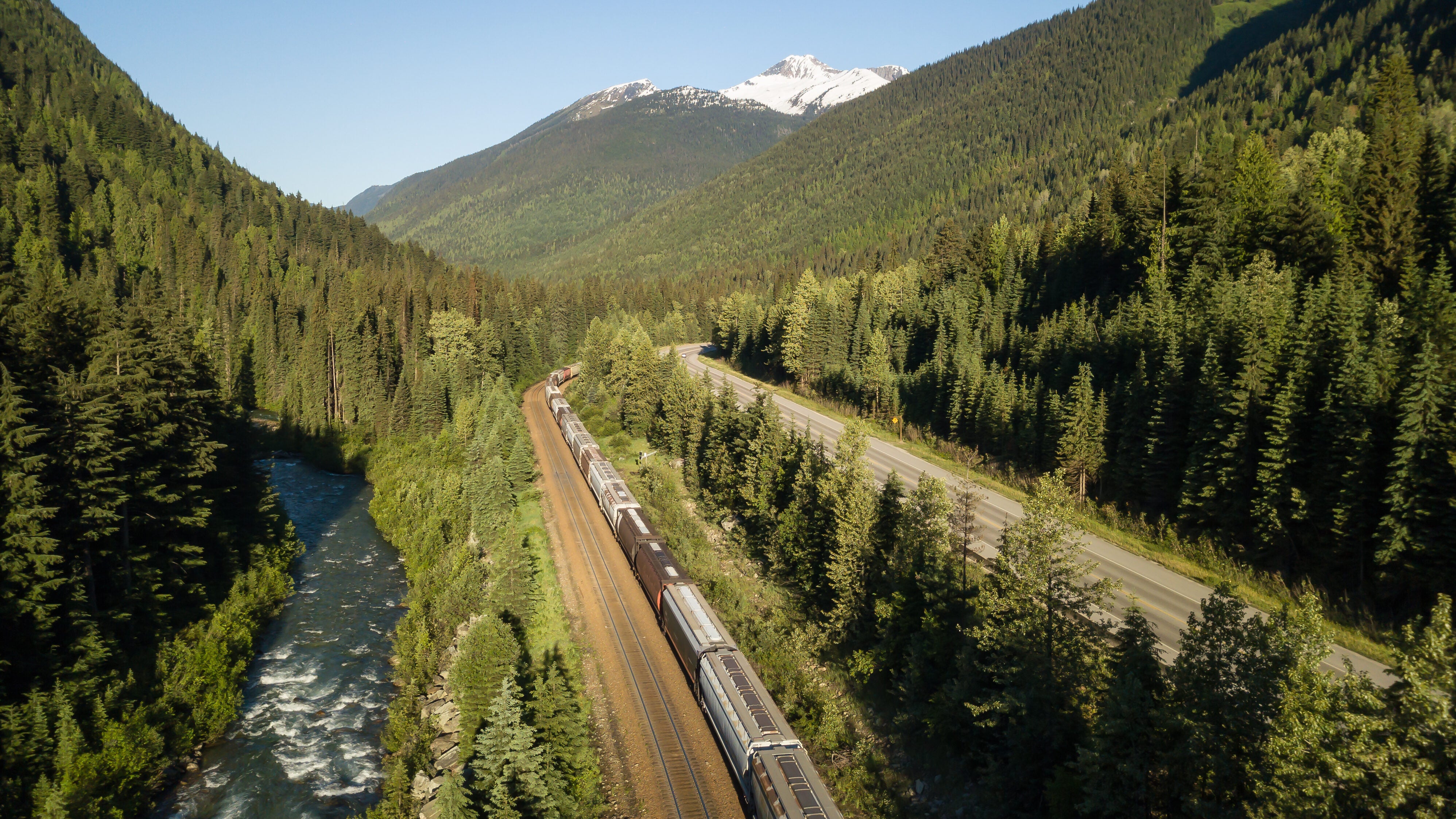 a train passes through a forest. There are mountains flanking both sides of the train, and there is a snow-capped mountain behind the train,