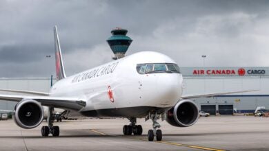 Frontal view of an Air Canada Cargo jet with cargo terminal in the background.