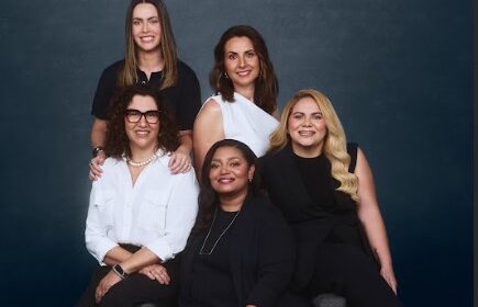 Na imagem do banner, em um fundo preto, iluminado no centro, temos cinco mulheres lado a lado, sorrindo e posando para a câmera. Da esquerda para a direita, a primeira mulher está em pé. Ela é branca de olhos castanho-escuros e cabelos lisos e castanhos, veste uma camiseta preta e usa um relógio preto no punho esquerdo. Na frente dela, temos a segunda mulher, que está sentada. Ela é uma mulher madura, branca, de olhos castanho-escuros e cabelos encaracolados pretos, na altura dos ombros. Ela veste uma camisa branca e uma calça preta, usa um colar de pérolas brancas, um relógio no punho esquerdo e óculos de grau de armação marrom. Em pé, ao lado da primeira mulher, temos a terceira. Ela é branca de olhos e cabelos castanho-escuros. Ela veste uma regata branca de um ombro só. Na frente dela, sentada, temos a quarta mulher. Ela é negra de olhos castanho-escuros e cabelos lisos e pretos. Ela veste uma blusa preta sobreposta por um cardigan preto e usa um colar dourado. Ao lado dela, temos a última. Ela é uma mulher branca de olhos castanho-escuros e cabelos loiros ondulados. Ela veste uma blusa preta sobreposta por um colete preto.