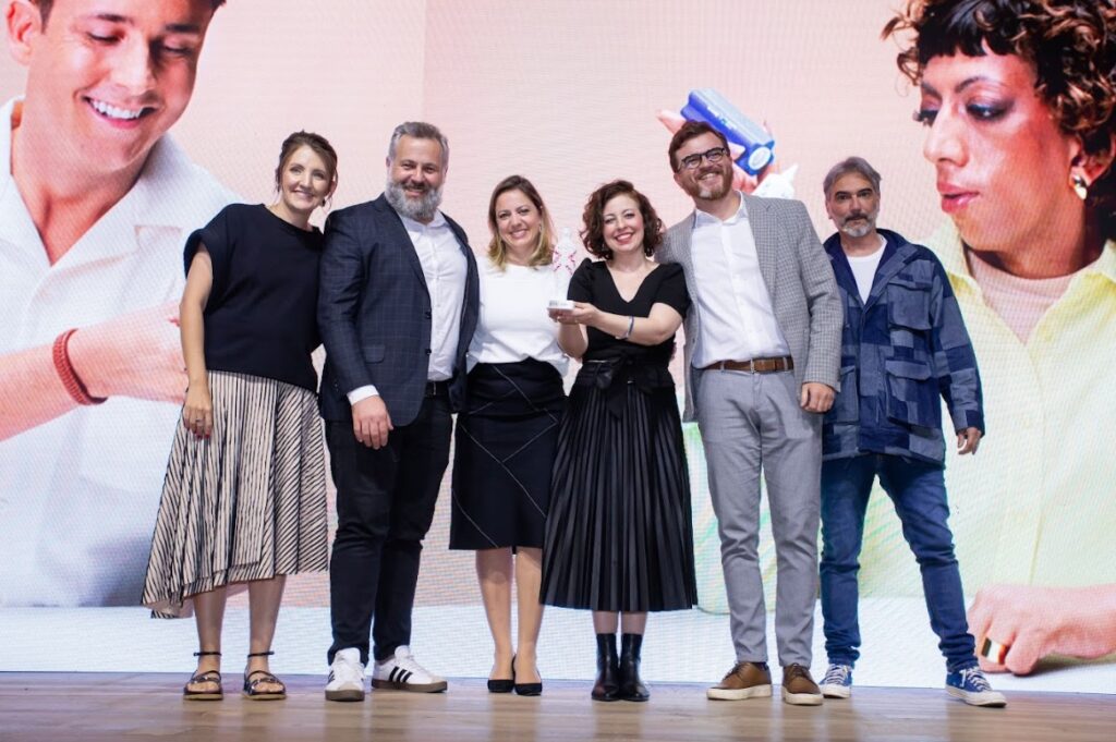 Imagem de seis pessoas em cima de um palco, são três mulheres e três homens, sendo uma das mulheres segura um troféu. Ao fundo, no telão, está projetada uma imagem de duas pessoas com deficiência, um homem e uma mulher, utilizando os produtos desenvolvidos pelo Grupo Boticário em parceria com a Mercur.