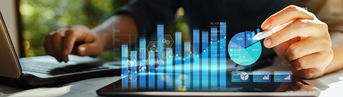 Cropped view of person at desk working with a laptop and tablet, with the screen containing charts and graphs overlaying the tablet