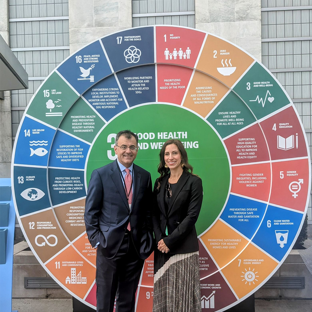 A man and a woman, smiling at the camera and standing in front of a large wheel that shows each of the United Nations’ sustainable development goals.