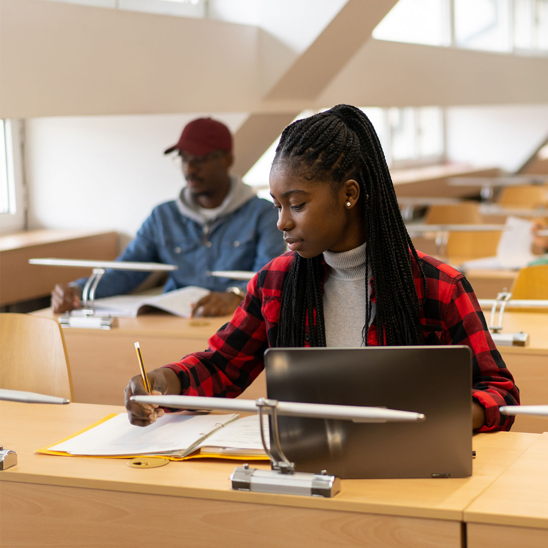 Students sitting at rows of tables with books and laptops, reading and writing