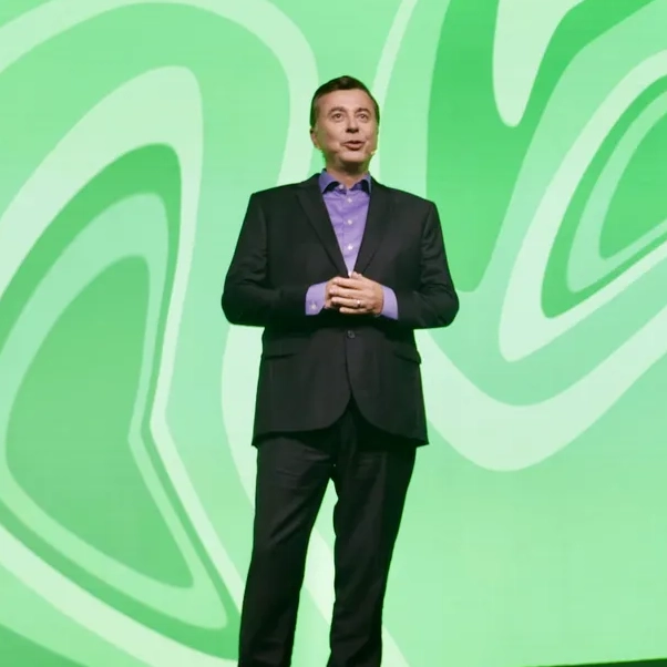 A man in a dark suit and blue shirt speaks on stage with a green abstract background.