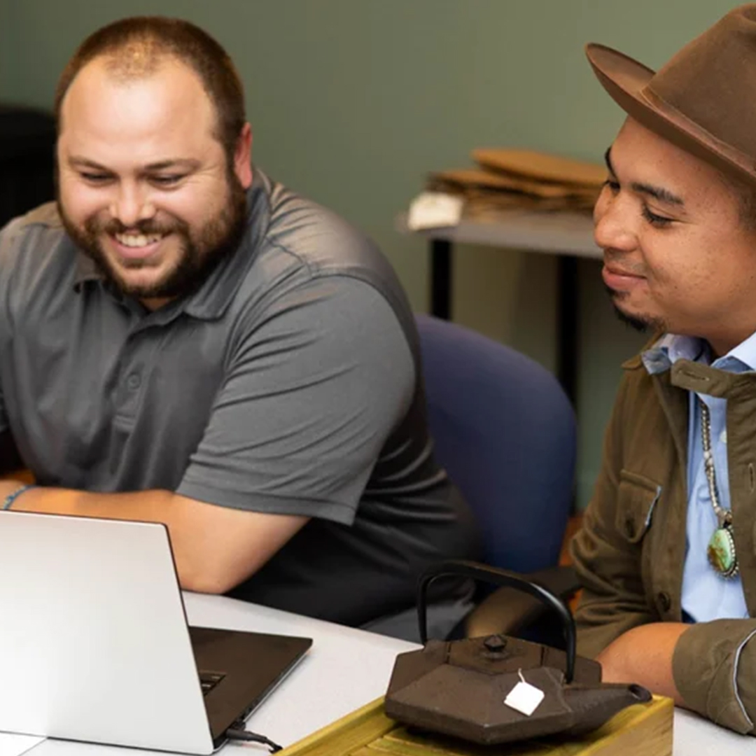 Two men smiling and sitting at a table looking at a laptop screen