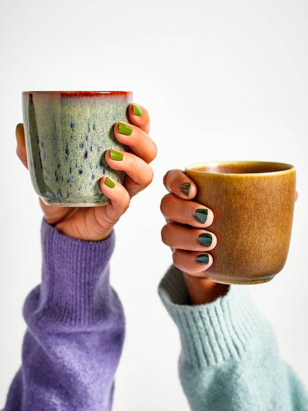 Two hands close to each other are holding two GLADELIG mugs in mixed colors against a white background.