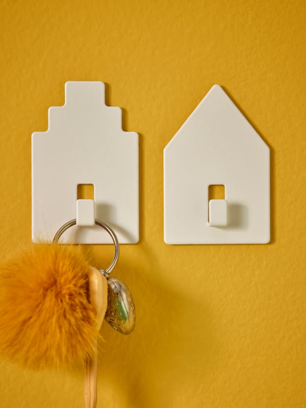 A wall section with two house-shaped TIPPVAGN self-adhesive hooks, arranged close to each other, one holding a keyring.