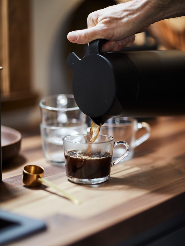 A person is pouring coffee from a UNDERLÄTTA vacuum flask into a IKEA 365+ mug, placed on a wooden surface.