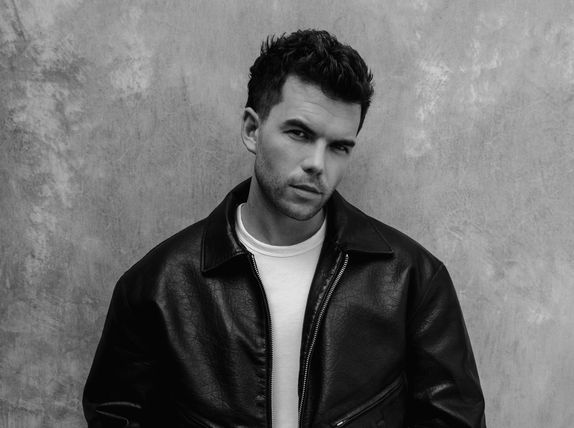 Luke Newton in a leather jacket leaning against a wall looking at the camera