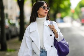 Person with short hair wears a white turtleneck with a white coat and purple bag