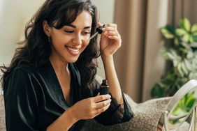 Smiling brunette person in black robe applies a serum to their cheek