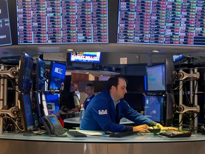 A trader leans over a desk while working on the floor of the New York Stock Exchange, surrounded by several computer screens. 