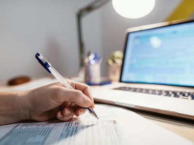 Close up of a hand holding a pen filling out tax forms with a laptop in the background