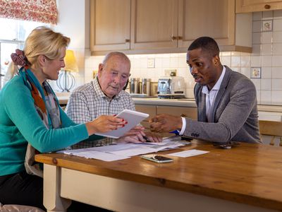 A financial advisor sits at a kitchen table with a senior investor and mature child and explains how they can contribute a gift of funds to an IRA account.