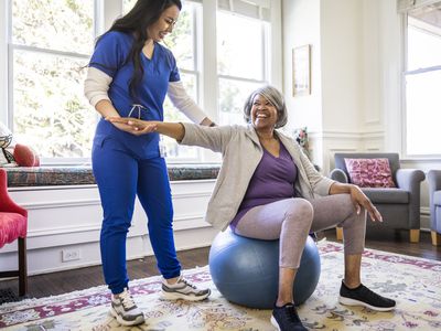 Gray-haired woman sits on a therapy ball while a physical therapist holds her arm up