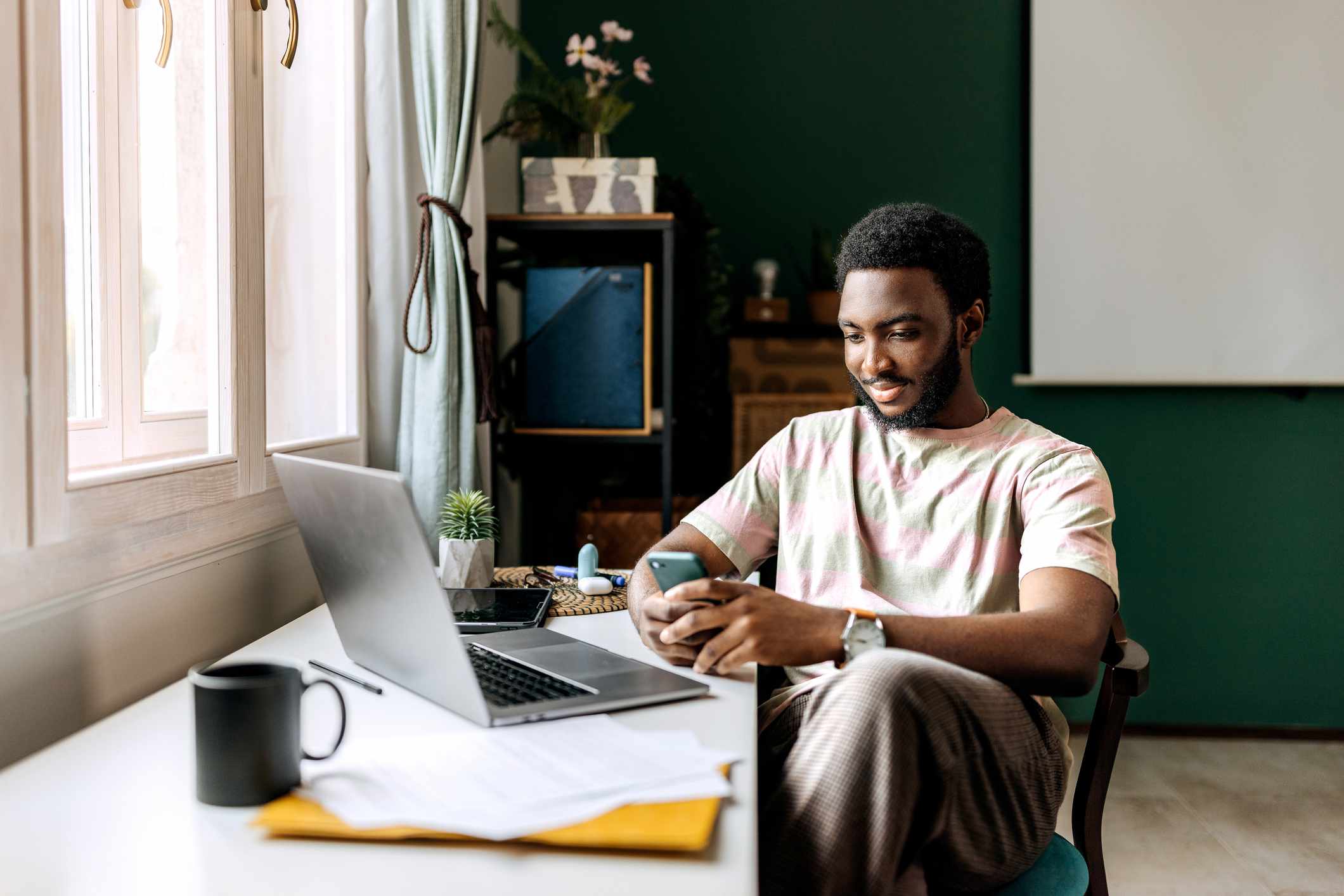 Young adult with beard reviews stock investments on phone while sitting next to a window.