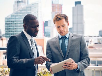 two men in suits looking at a tablet outside with a skyline in the background