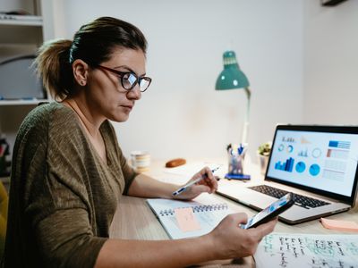 Woman looking at market charts on her smartphone and a laptop, seated at a desk