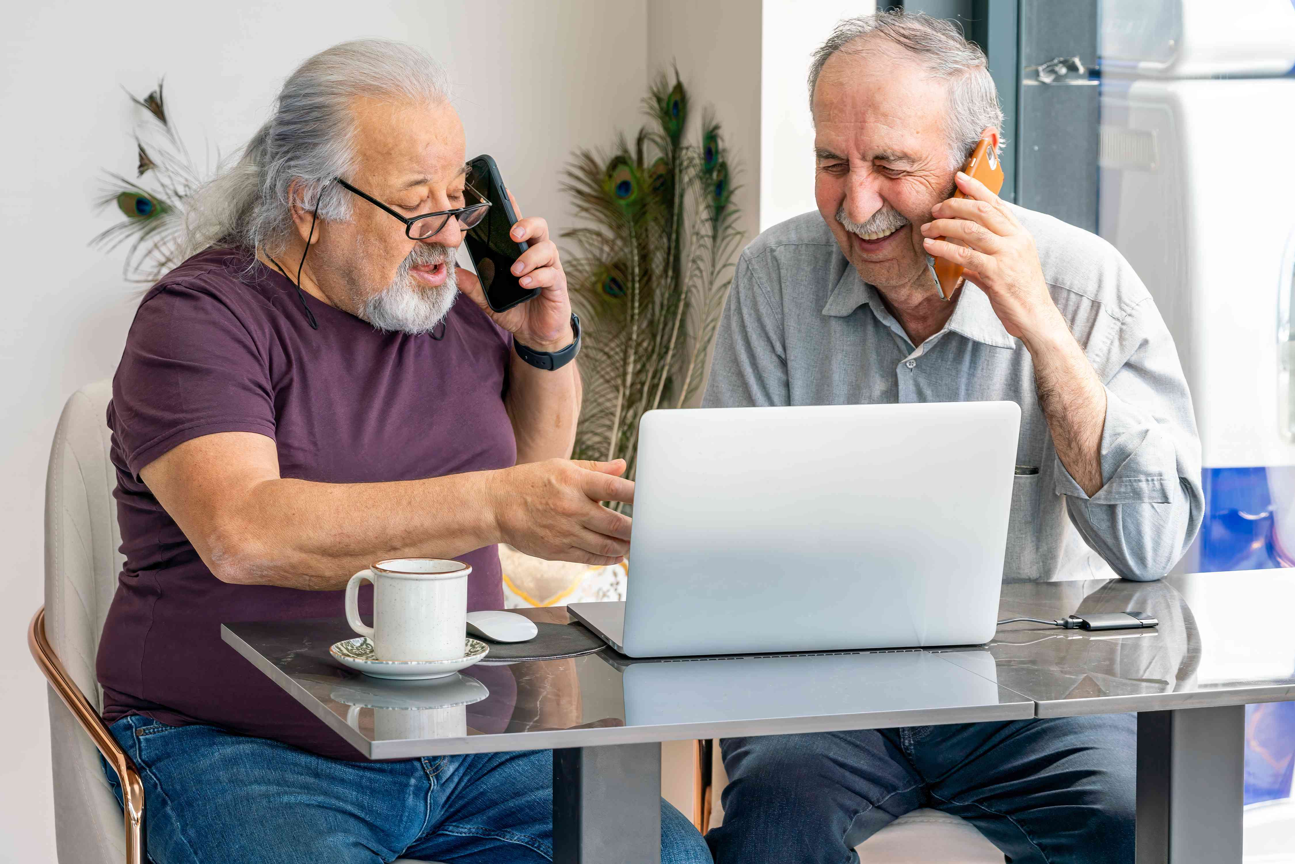 Two men look at a laptop, and both are on their mobile phones.