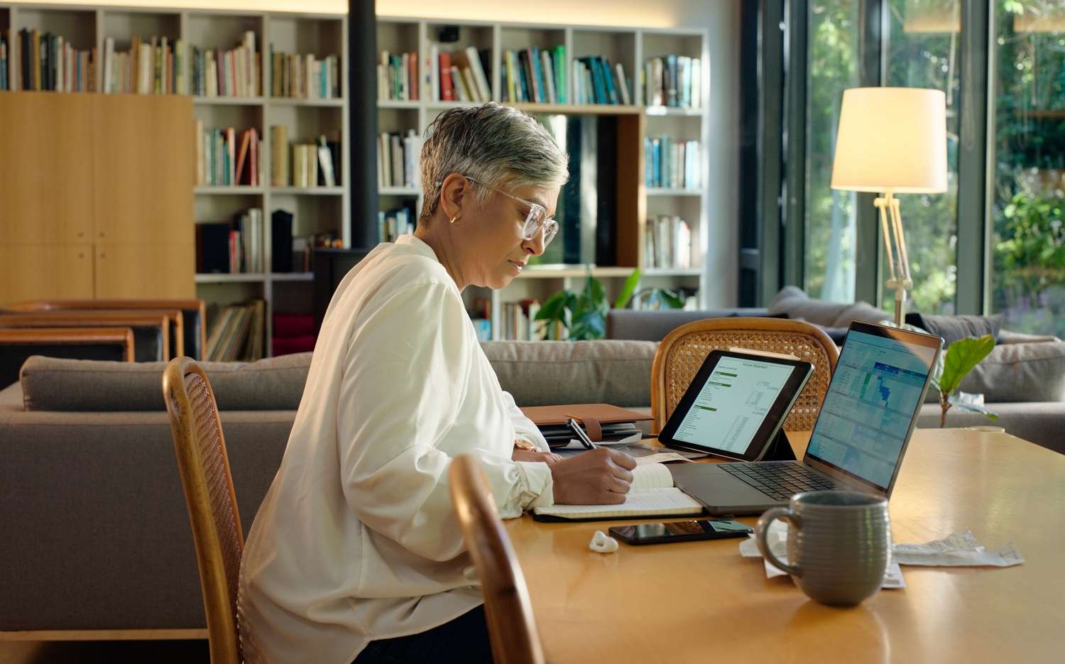 older woman takes notes while working on laptop and tablet with charts on screen