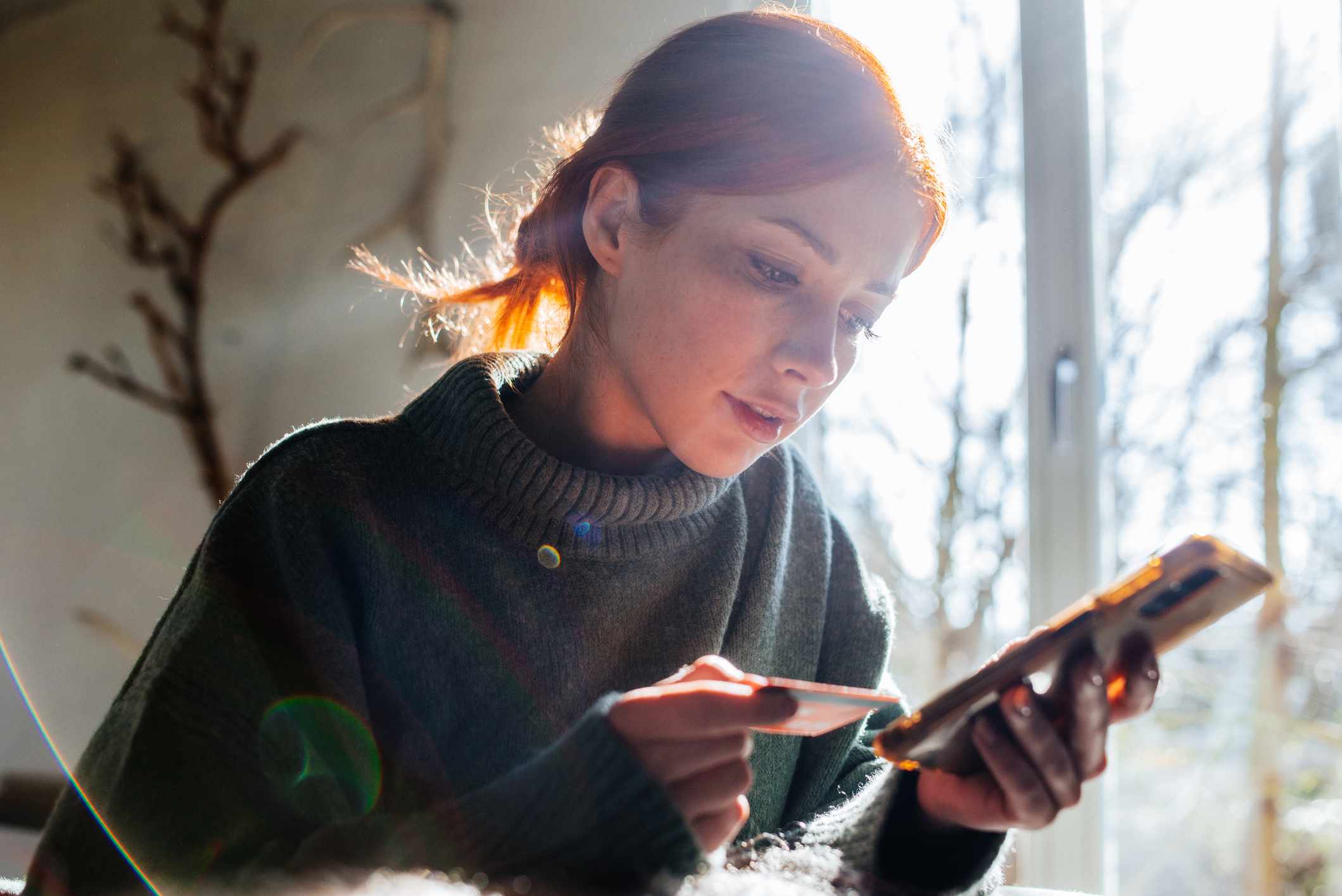 A young woman is sitting near a sunny window at home, looking carefully at both her smartphone and a credit card