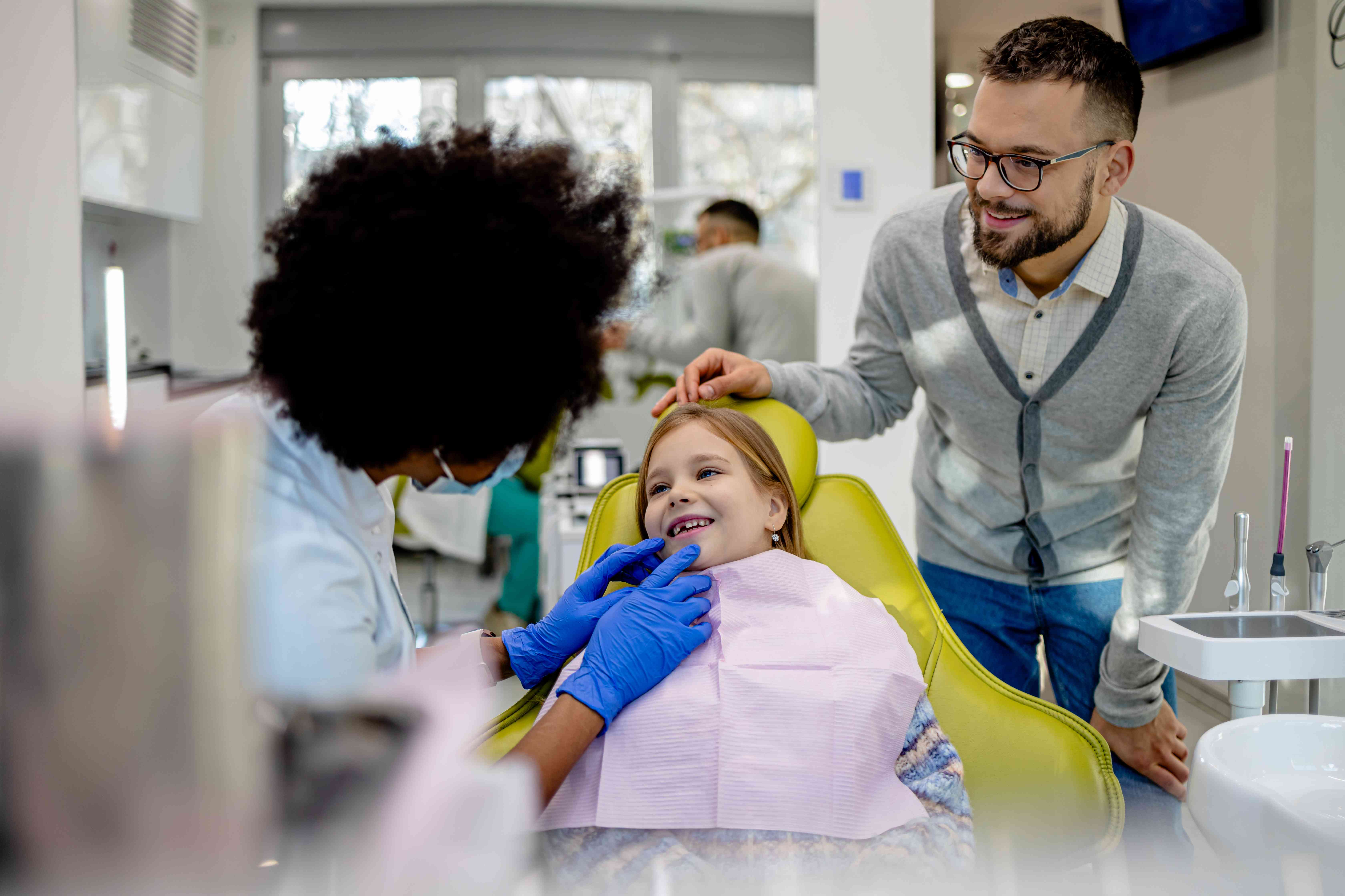 A dentist examines a young girl's teeth as her father looks on