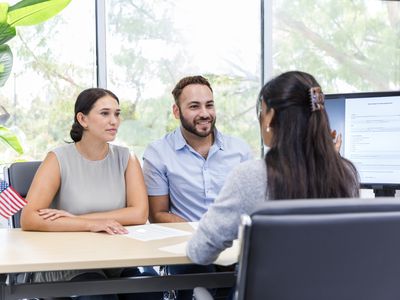 A man and a woman sit at a desk in a bank office talking with a female banker. There's a computer screen with a document on it.