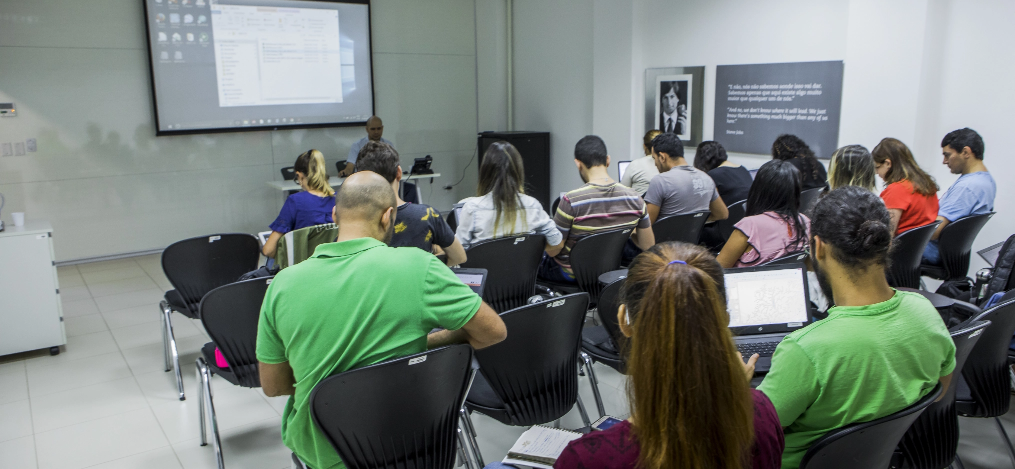 Sala de aula com 15 estudantes de costas para câmera olhando para o professor que está sentado de frente para eles. Atrás do professor, há um telão.