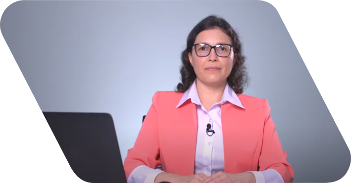 Mulher de cabelo médio vestindo uma camisa de botão branca com um terno rosa. Ela fala para câmera em frente a um fundo cinza.