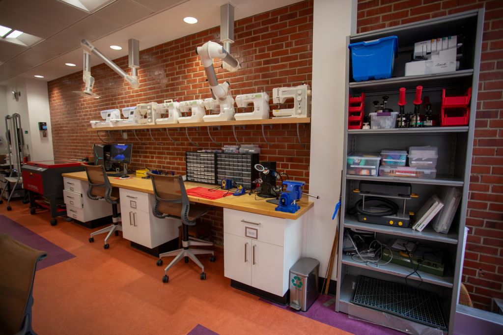 Work tables with a row of sewing machines and shelves containing button makers and crafting supplies