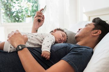 Father lying with his baby on his chest in bed while checking a sleep app on his iPhone