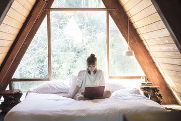 A stock photo of a woman sitting on bed wearing headphones around her neck and staring at Windows 10 Surface laptop.