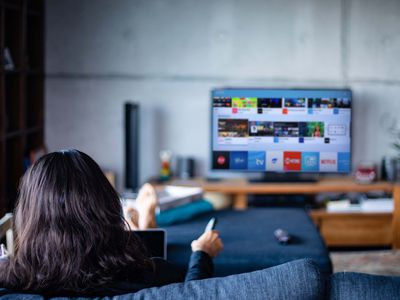 A person sitting on a couch usign their TV's remote to navigate the Samsung App Store on a TV. The TV is far away and appears to have a lot of apps from the Samsung App store on the screen.